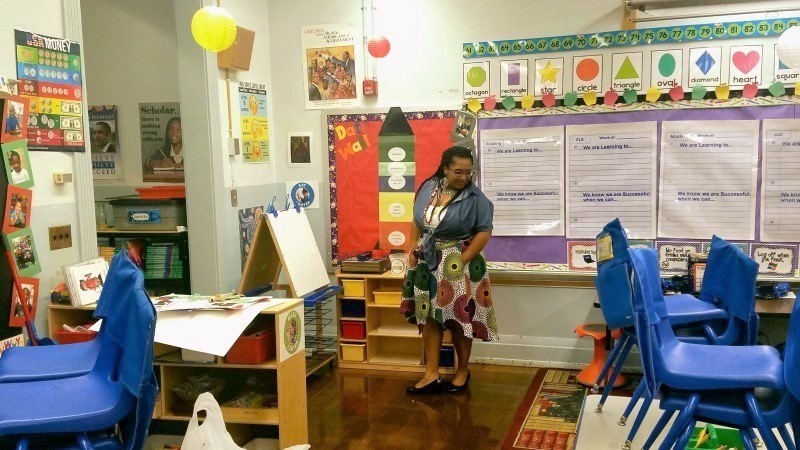 A teacher is standing in front of different activities on bulletins boards, and there are a few stacks of blue chairs.