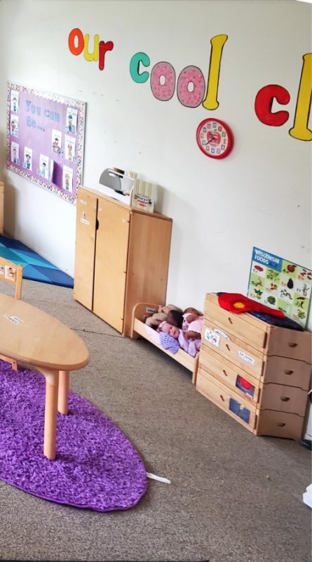 Play area that includes some furniture, a clock on the wall, a bulletin board with the title “You can be…”, and a bed with baby dolls in it.